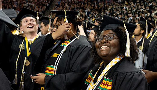 Students in full undergraduate graduation regalia celebrate at commencement ceremony in McCamish Pavilion. One student is raising their arms in celebration, another is arranging their tassel, and the third is smiling while looking upwards. The crowd of graduates is visible in the background.