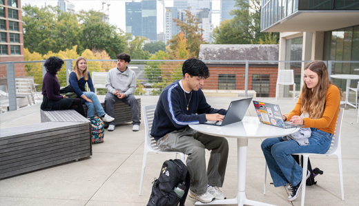 A group of students study on a concrete patio area outside of Georgia Tech’s library. Students on benches and sitting at tables take up the foreground, and the Atlanta skyline is visible in the background.